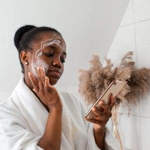 dark skinned woman cleansing her face and looking at her phone