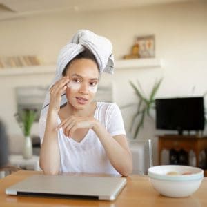 woman applying eye mask, sitting at a table in front of a laptop