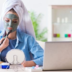 woman applying a green face mask whilst watching a tutorial on a laptop